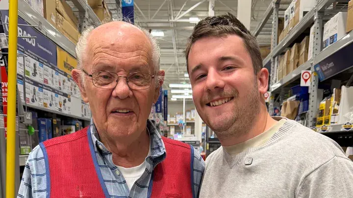 Les, an 83-year-old man in glasses, smiling beside a younger man at the store where he works